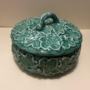 Puede incluir: Un recipiente redondo de cerámica verde turquesa con tapa. La tapa presenta un asa curva y está adornada con un diseño detallado de hojas y uvas. El cuerpo del recipiente también muestra un patrón de hojas en relieve.