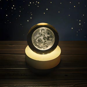 May include: A glowing moon lamp with a detailed lunar surface, set within a dark frame. The lamp sits on a round, illuminated base, creating a warm, ambient light. The background features a dark blue sky with scattered stars, evoking a night sky theme.