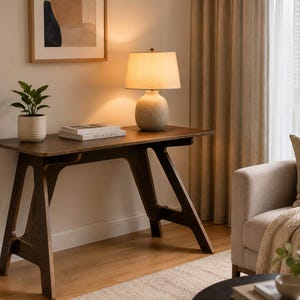 May include: A dark brown wooden desk with an A-frame base, featuring a light-coloured ceramic table lamp with a fabric shade, a small potted green plant in a ribbed white pot, and a stack of two books placed on the flat rectangular surface.
