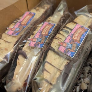 Könnte beinhalten: Verpackte Biscotti mit Schokosauce, beschriftet mit "Gnome Baked Goods". Die Biscotti befinden sich in durchsichtigem Kunststoff, angeordnet in einem Pappkarton. Die Etiketten zeigen eine Zwerg-Illustration mit rosa und blauem Hintergrund.