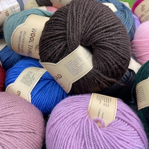 K&ouml;nnte beinhalten: Eine Ansammlung runder Wollkn&auml;uel in verschiedenen Farben wie Dunkelbraun, leuchtendem Blau, hellem Lila, Altrosa und T&uuml;rkis. Jedes Kn&auml;uel hat ein beiges Papieretikett mit dem Text Wool Thread, Delicate and Soft, Intimate Warmth.