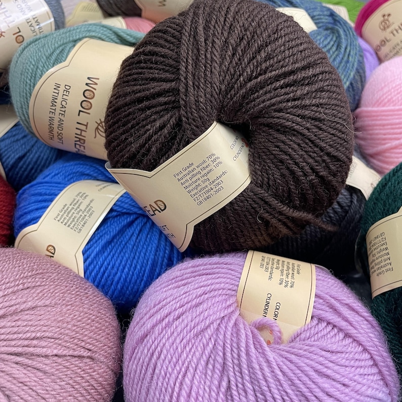 K&ouml;nnte beinhalten: Eine Ansammlung runder Wollkn&auml;uel in verschiedenen Farben wie Dunkelbraun, leuchtendem Blau, hellem Lila, Altrosa und T&uuml;rkis. Jedes Kn&auml;uel hat ein beiges Papieretikett mit dem Text Wool Thread, Delicate and Soft, Intimate Warmth.
