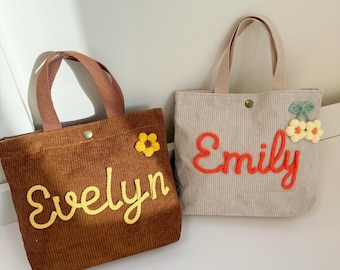 Könnte beinhalten: Zwei Cord-Tragetaschen, eine braune und eine beige, mit gestickten Namen und floralen Akzenten. Die braune Tasche hat "Evelyn" in gelber Schrift, und die beige Tasche hat "Emily" in roter Schrift. Beide haben passende Griffe.