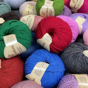K&ouml;nnte beinhalten: Eine Sammlung mehrfarbiger Wollkn&auml;uel, jeweils umwickelt mit einem beigen Papieretikett mit dem Text Wool Thread, Delicate and Soft, Intimate Warmth. Die Farben umfassen Rot, Blau, Gr&uuml;n, Rosa, Violett, Grau und Braun in verschiedenen Texturen.