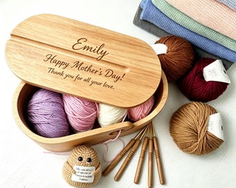 Könnte beinhalten: Holzkiste zur Garnaufbewahrung mit dem Namen "Emily" und der Aufschrift "Happy Mother's Day!". Sie enthält Garnknäuel in Lila, Rosa und Weiß. Häkelnadeln und weitere Garnknäuel sind ebenfalls zu sehen.