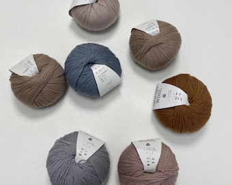 Könnte beinhalten: Sieben Knäuel Merinowolle in verschiedenen Farben, darunter Braun, Blau und Rosa. Jedes Knäuel hat ein Etikett. Das Garn ist in einem kreisförmigen Muster auf einer weißen Oberfläche angeordnet, geeignet für Strick- oder Häkelprojekte.