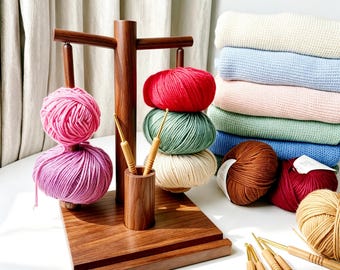 Könnte beinhalten: Ein hölzerner Garnhalter mit rosa, roten, grünen und cremefarbenen Garnknäueln. Häkelnadeln befinden sich in einem Holzhalter. Im Hintergrund ein Stapel gefalteter Pullover in verschiedenen Pastellfarben. Zusätzliche Garnknäuel sind ebenfalls sichtbar.