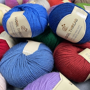 K&ouml;nnte beinhalten: Eine Ansammlung runder Wollkn&auml;uel in verschiedenen Farben, darunter leuchtendes Blau, Dunkelblau, Rot und Violett. Jedes Kn&auml;uel hat ein beiges Papieretikett mit dem Text WOOL THREAD, DELICATE AND SOFT, INTIMATE WARMTH und einem Symbol f&uuml;r Stricknadeln.