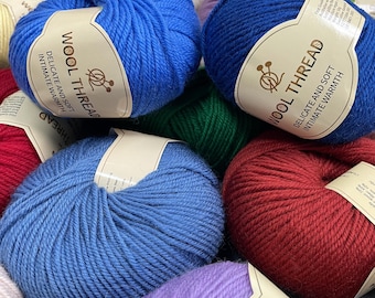 Puede incluir: Una colección de ovillos de lana redondos en varios colores, incluyendo azul brillante, azul oscuro, rojo y morado. Cada ovillo tiene una etiqueta de papel beige con el texto WOOL THREAD, DELICATE AND SOFT, INTIMATE WARMTH y un icono de agujas.