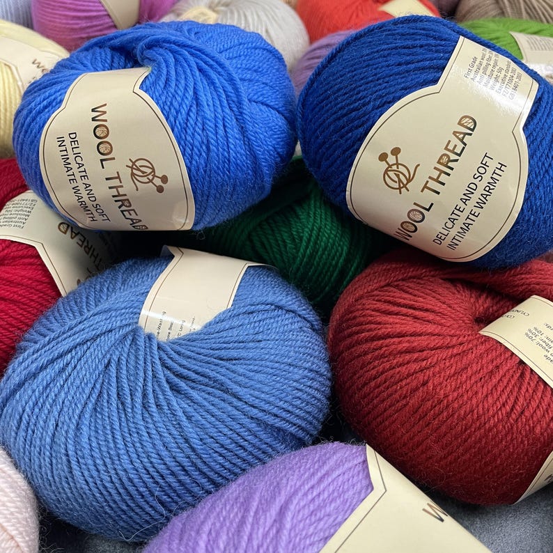 K&ouml;nnte beinhalten: Eine Ansammlung runder Wollkn&auml;uel in verschiedenen Farben, darunter leuchtendes Blau, Dunkelblau, Rot und Violett. Jedes Kn&auml;uel hat ein beiges Papieretikett mit dem Text WOOL THREAD, DELICATE AND SOFT, INTIMATE WARMTH und einem Symbol f&uuml;r Stricknadeln.