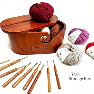 Puede incluir: Una caja de almacenamiento de hilo de madera con tapa, personalizada con el nombre "Emily". La caja está llena de varios colores de hilo y rodeada de ganchillos y madejas de lana. El texto "Yarn Storage Box" es visible.