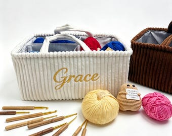 Könnte beinhalten: Ein weißer Korb aus Kordstoff mit dem in Gold gestickten Namen Grace, ausgestattet mit Inneneinteilungen und Griffen. Der Korb enthält Garnknäuel und ist umgeben von Häkelnadeln aus Holz sowie einem gelben und einem pinken Garnknäuel.