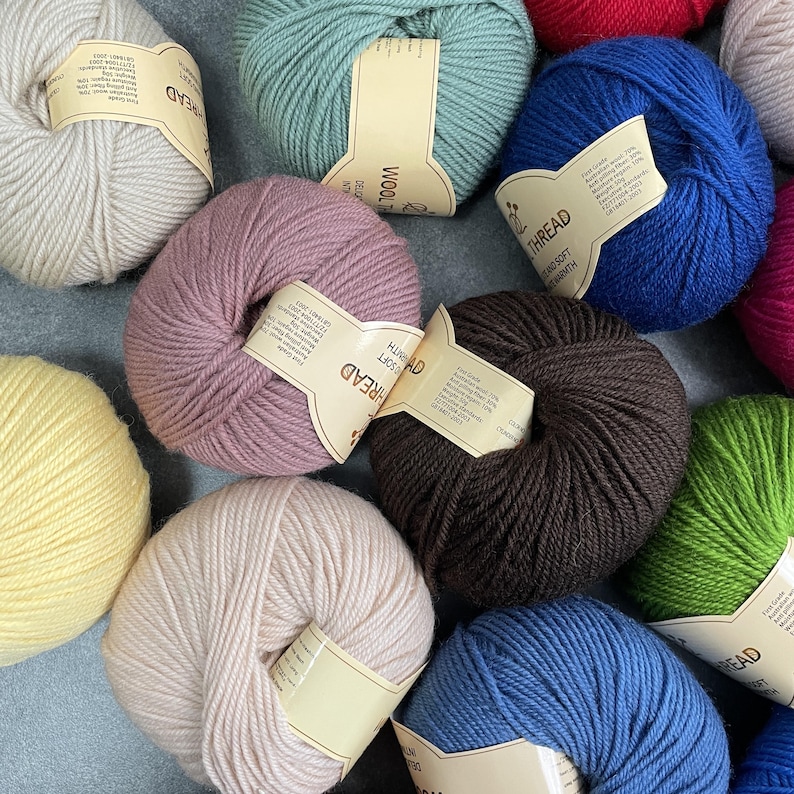 K&ouml;nnte beinhalten: Eine Sammlung runder Garnkn&auml;uel in verschiedenen Farben, darunter Creme, Hellgr&uuml;n, K&ouml;nigsblau, Mauve, Dunkelbraun, Gelb und Hellgr&uuml;n. Jeder Kn&auml;uel hat ein beiges Papieretikett mit dem Text Wool Thread und weiteren Produktdetails.
