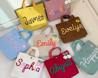 Könnte beinhalten: Eine Sammlung von Cord-Tragetaschen in verschiedenen Farben, jede mit einem Namen in einer Kontrastfarbe personalisiert. Die Taschen haben passende Griffe und florale Verzierungen. Namen sind James, Amelia, Evelyn, Emma, Emily, Sophia, Abigail und Harper.