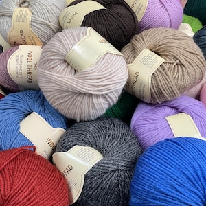 K&ouml;nnte beinhalten: Eine Ansammlung runder Wollkn&auml;uel in verschiedenen Farben wie Rot, Blau, Grau, Braun, Beige und Lavendel. Jedes Kn&auml;uel hat ein beiges Papieretikett mit dem Aufdruck Wool Thread, Delicate and Soft, Intimate Warmth und der Gewichtsangabe 50g.