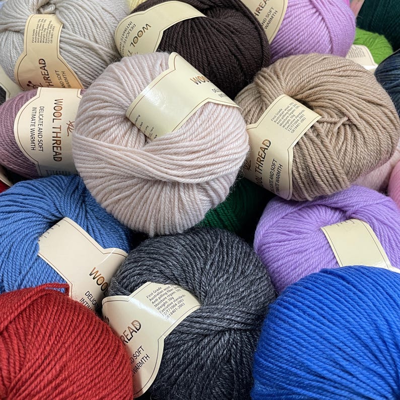 K&ouml;nnte beinhalten: Eine Ansammlung runder Wollkn&auml;uel in verschiedenen Farben wie Rot, Blau, Grau, Braun, Beige und Lavendel. Jedes Kn&auml;uel hat ein beiges Papieretikett mit dem Aufdruck Wool Thread, Delicate and Soft, Intimate Warmth und der Gewichtsangabe 50g.