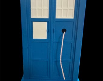 Puede incluir: Una maqueta de cabina de policía azul con ventanas blancas y las palabras "POLICE BOX" en la parte superior. También son visibles las palabras "PUBLIC CALL". Un cordón blanco cuelga de la parte delantera.