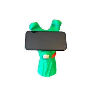 Puede incluir: Un soporte para teléfono de mano verde con una banda roja alrededor de la muñeca, sosteniendo un teléfono inteligente negro con una pantalla en blanco.