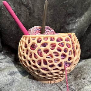 May include: A pink yarn bowl with a multi-colored ball of yarn inside. The bowl has a lattice design and the words "SHIRL SP" are printed on the bottom. A pink crochet hook and a gray knitting needle are sticking out of the yarn.