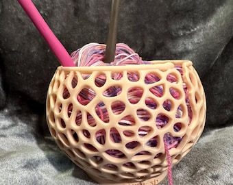 May include: A pink yarn bowl with a multi-colored ball of yarn inside. The bowl has a lattice design and the words "SHIRL SP" are printed on the bottom. A pink crochet hook and a gray knitting needle are sticking out of the yarn.