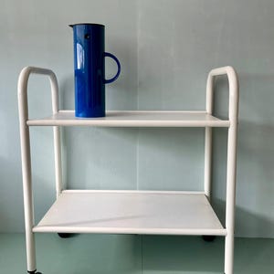 Op de afbeelding: Een witte metalen serveerwagen met twee geperforeerde planken en gebogen handgrepen. Een kobaltblauwe kan met een zwart deksel en handvat staat op de bovenste plank. De wagen heeft vier zwarte wielen.