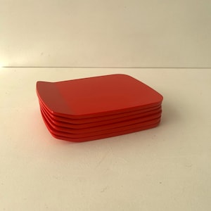 Könnte beinhalten: Ein Stapel aus sechs quadratischen roten Tellern mit abgerundeten Ecken und einem leicht nach oben gebogenen Rand an einer Seite. Die Teller sind in Farbe und Form einheitlich und haben eine glatte, matte Oberfläche. Der Stapel ist ordentlich angeordnet und zeigt das konsistente Profil jedes einzelnen Tellers.