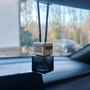 May include: A car air freshener with a wooden top and a black glass bottom. The wooden top has the text "VIOLENT LITTLE THING" and a dragon graphic. The air freshener hangs from a black cord.