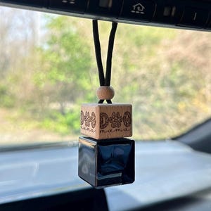 May include: A car air freshener hanging from a rearview mirror, featuring a light wood cube with the engraved text DOG MAMA and paw prints, topped with a small wooden bead, and attached to a black glass cube base by a black cord.