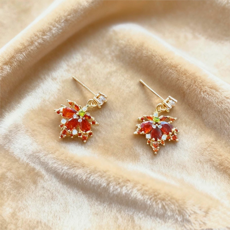 May include: A pair of gold-colored drop earrings. Each earring features a maple leaf design with red and white gemstones, accented with a small green stone in the center. The earrings are set against a soft, beige background.