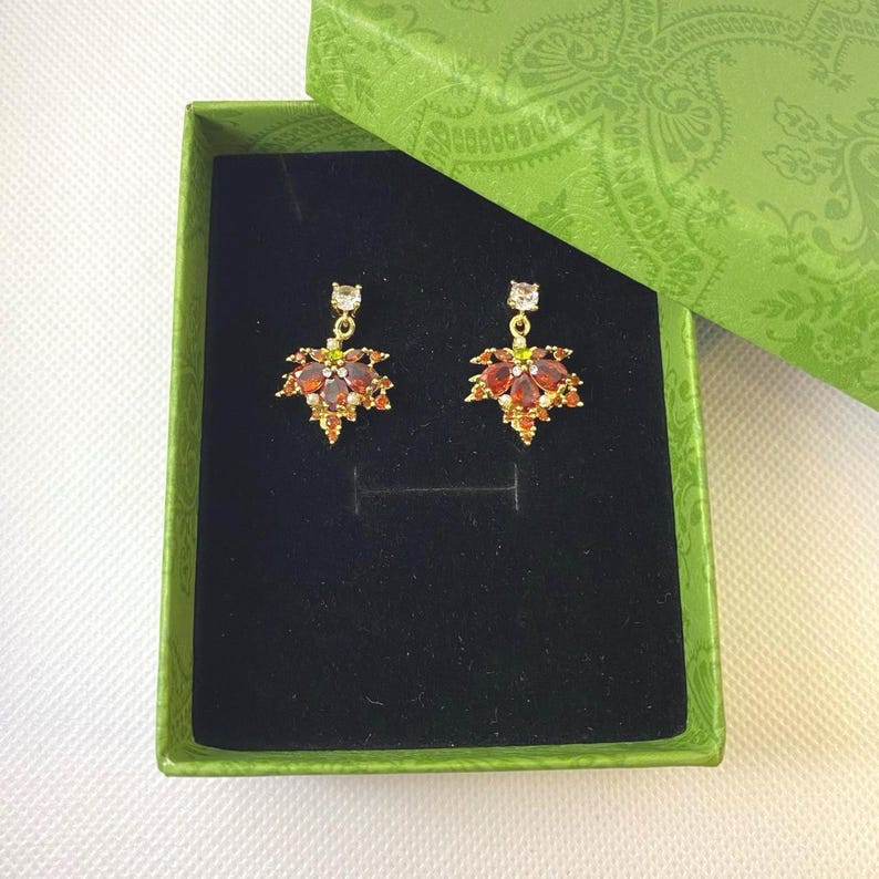 May include: A pair of gold-toned earrings with a leaf design, set with red and orange gemstones. The earrings are presented in a green box with a black interior. The box is open, revealing the jewelry.