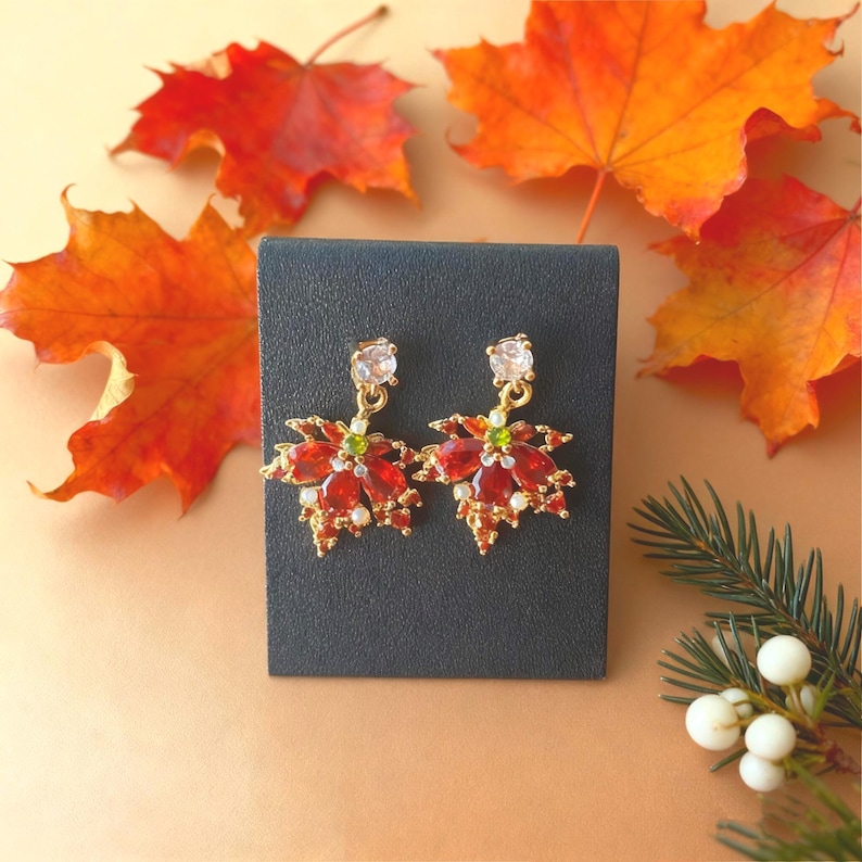 May include: A pair of gold-toned earrings shaped like maple leaves, adorned with red and clear gemstones. The earrings have a small green gemstone in the center and a clear gemstone at the top. The earrings are displayed on a black surface with autumn leaves in the background.