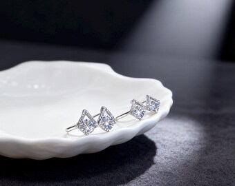 May include: Two pairs of silver-toned stud earrings with a geometric design. Each earring features a central, clear, square-cut stone, surrounded by smaller, clear stones. The earrings are displayed on a white, organic-shaped dish against a dark background.