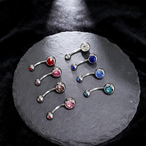 May include: A collection of curved belly button rings with various colored gemstones, including red, pink, blue, and clear. The jewelry is displayed on a dark gray, round stone surface against a black background.