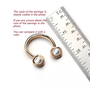 May include: Gold-toned, U-shaped earrings with a pearl at each end. A ruler is next to the earrings, with text that reads, "The style of the earrings is clearly visible in the photo. If you are unsure about the size of the earrings in the photo, you can compare it with a ruler."