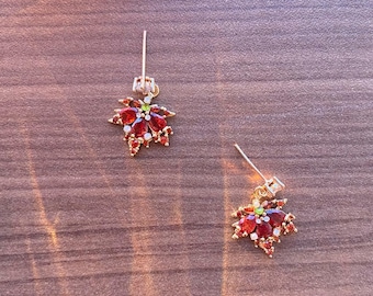 May include: A pair of gold-coloured earrings, each with a maple leaf design. The leaves are decorated with red and white gemstones, with a small green gem at the centre. The earrings are displayed on a wooden surface.