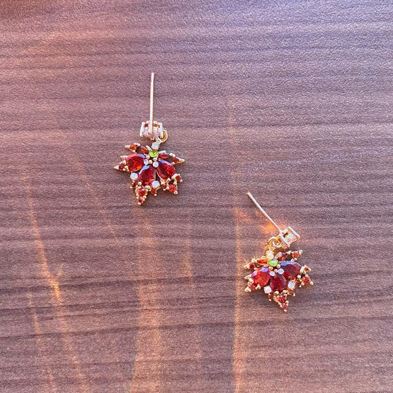 May include: A pair of gold-colored earrings, each featuring a maple leaf design. The leaves are adorned with red and white gemstones, with a small green gem at the center. The earrings are set against a wooden surface.