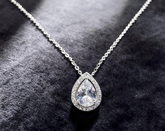 May include: A silver necklace with a pear-shaped pendant. The pendant is encrusted with small, clear stones around a larger, pear-cut stone. The chain is delicate and silver, and the necklace is set against a dark background.