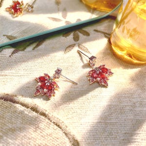 May include: A pair of gold-toned stud earrings featuring a maple leaf design. The earrings are adorned with red and pink gemstones, with a small square gem at the post. The earrings are set against a soft, patterned background.