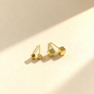 May include: Two pairs of gold stud earrings. One pair has a geometric, faceted design, the other a cube shape. The earrings are on a light beige surface, with shadows adding depth. The earrings are likely made of gold.