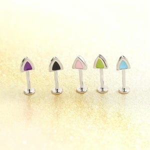 May include: Five silver-colored lip or ear studs with triangular tops in purple, black, pink, green, and blue. Each stud has a flat base and a post. The background is a light, textured gold.