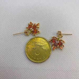 May include: A pair of gold-colored earrings with a floral design. Each earring features a cluster of orange and red gemstones, with a small green gemstone and a white pearl accent. A Canadian five-dollar coin is next to the earrings.