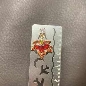 May include: A gold-toned earring with a red and gold floral design, accented with small white and green gems. The earring is approximately 1 inch in length, with a clear, round gem at the top.