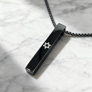 Peut inclure: Un collier pendentif noir avec un pendentif rectangulaire. Le pendentif présente une étoile de David blanche. Le collier a une chaîne sombre et est présenté sur une surface en marbre.