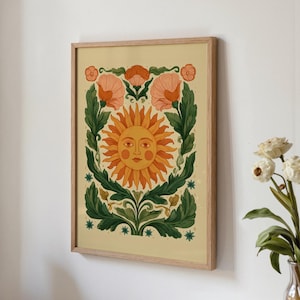 May include: A framed wall art print featuring a central sun with a human face, surrounded by large green leaves and orange flowers on a light yellow background. The illustration is enclosed in a light-colored wooden frame with a clean border.