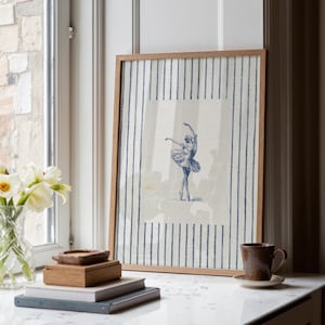 May include: A framed wall art print featuring a blue ink illustration of a ballerina in a tutu performing a dance pose, centered on a cream-colored background with vertical blue pinstripes, enclosed in a simple light brown wooden frame.