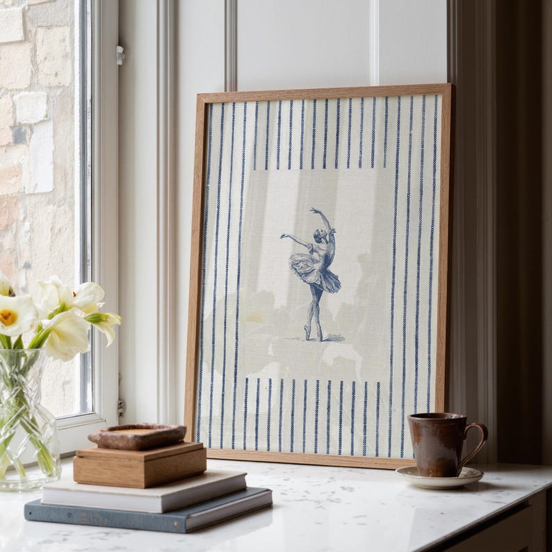 May include: A framed wall art print featuring a blue ink illustration of a ballerina in a tutu performing a dance pose, centered on a cream-colored background with vertical blue pinstripes, enclosed in a simple light brown wooden frame.