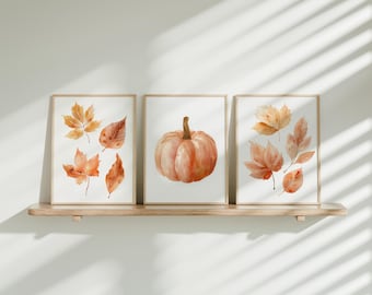 May include: Three framed watercolor prints featuring autumn themes. The prints depict orange and yellow leaves and a pumpkin. The frames are light-colored, and the prints are displayed on a wooden shelf against a white wall.