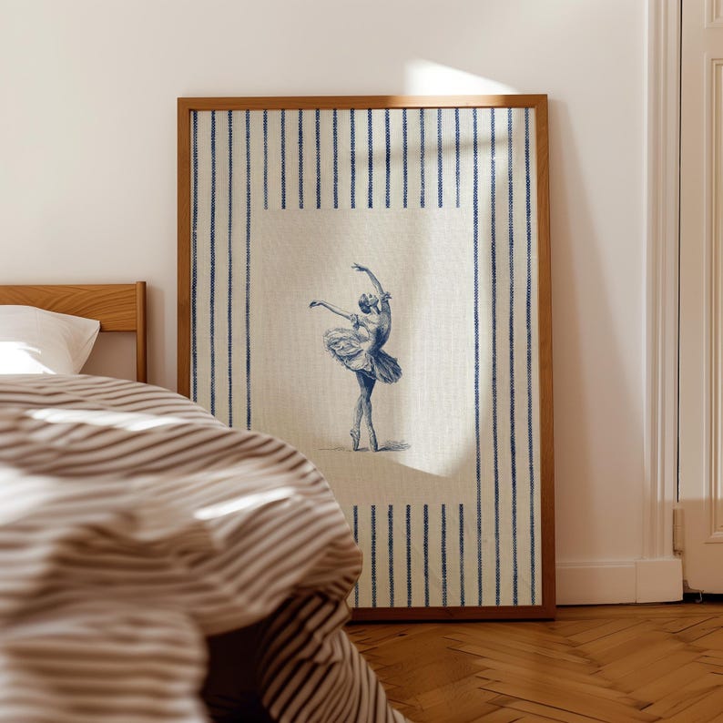 May include: A framed wall art print featuring a blue ink illustration of a ballet dancer in a tutu on a cream-colored background. The artwork is bordered by a pattern of vertical blue stripes on a cream fabric-like texture within a wooden frame.