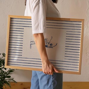 May include: A rectangular framed artwork featuring a light-colored fabric background with horizontal blue stripes along the left and right edges. In the center, a blue ink illustration depicts a figure in a dynamic, reclining pose.