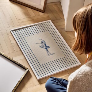 May include: A rectangular framed wall art piece featuring a centered blue ink illustration of a ballet dancer in a tutu. The background of the print consists of vertical blue stripes on a cream-colored field, enclosed in a simple white frame.